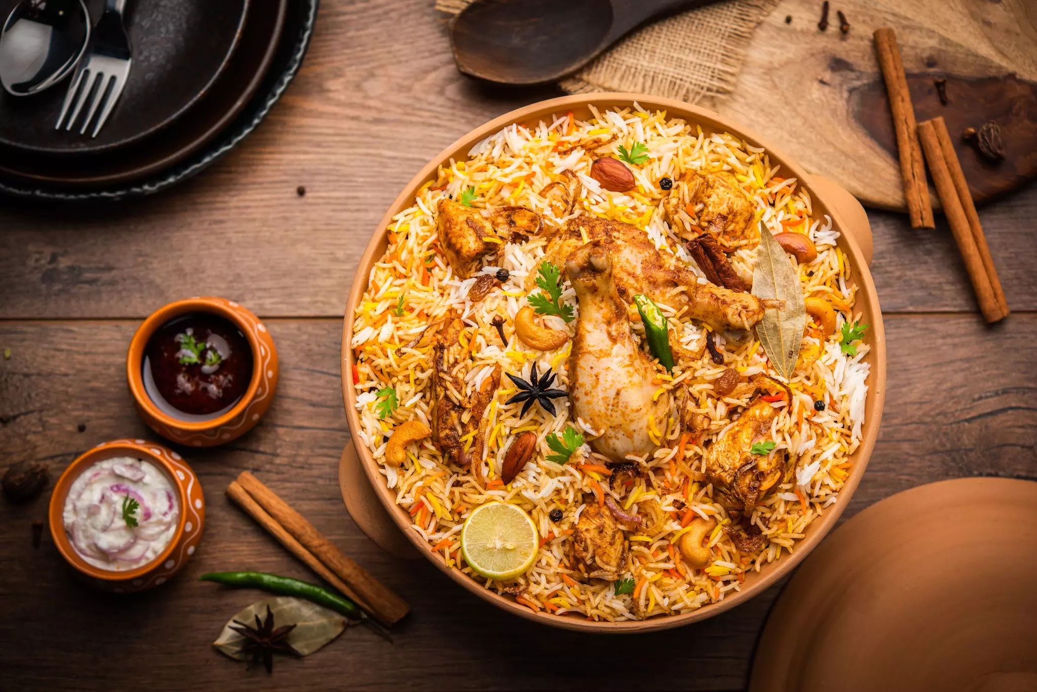 Chicken biryani in a round clay pot with smaller sauce pots and cinnamon sticks on table