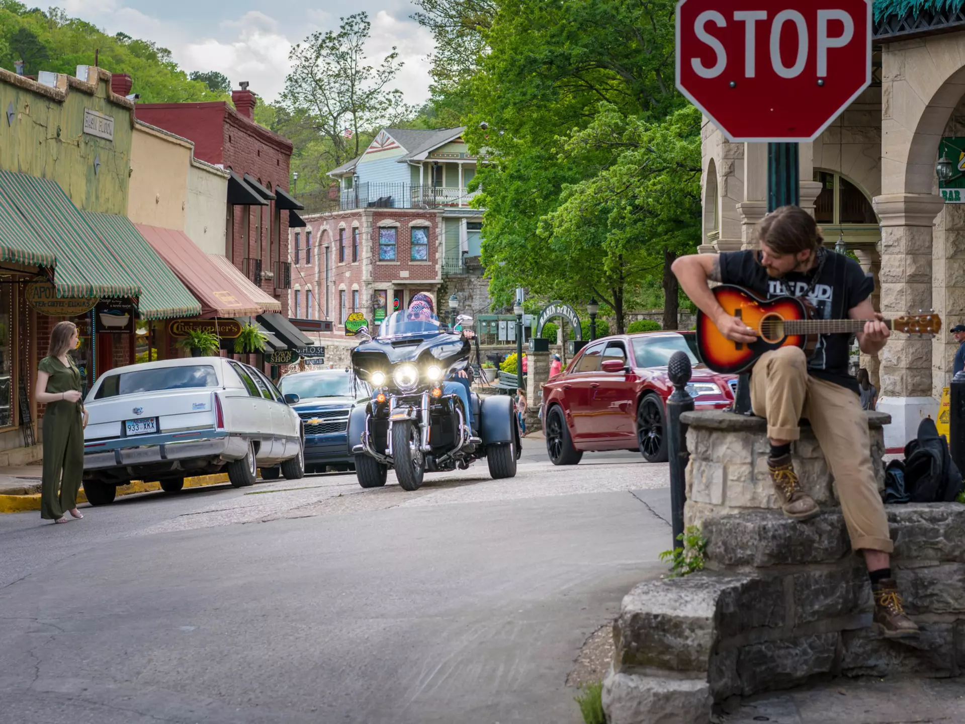 A biker in a three-wheel vehicle drives down the street of a town. A musician with a guitar is seen on a porch to the right.