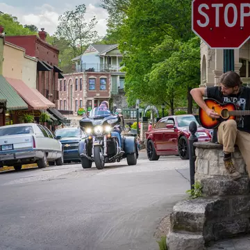 A biker in a three-wheel vehicle drives down the street of a town. A musician with a guitar is seen on a porch to the right.
