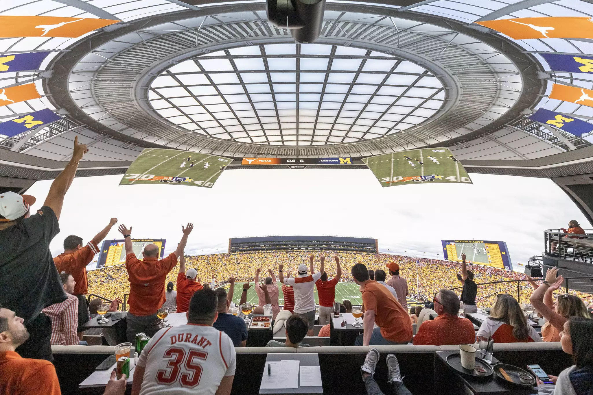 Fans sitting and standing, some with arms above their heads cheering, as a football game appears on a curved screen that gives the illusion of being in the stadium.
