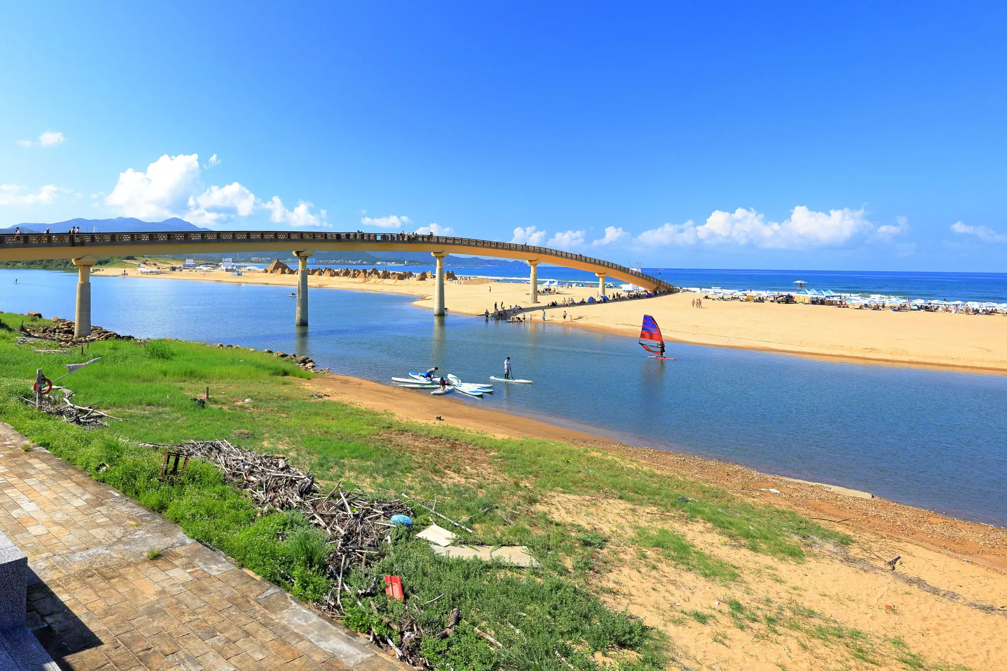 A river mouth with golden sandy beaches either side. People paddleboard and sail in the calm waters.