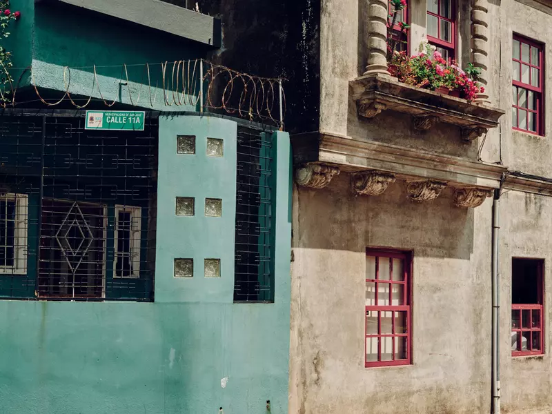The exterior of two buildings on a San Jose, Costa Rica block. One is blue with some barbed wire above the first floor, one is white with a window box of flowers and orange window trim.. 