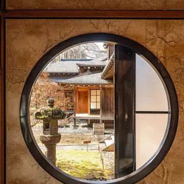 Wooden villa wings, stone lantern and mossy garden seen through a circular window at Tamozawa Imperial Villa, Nikko
