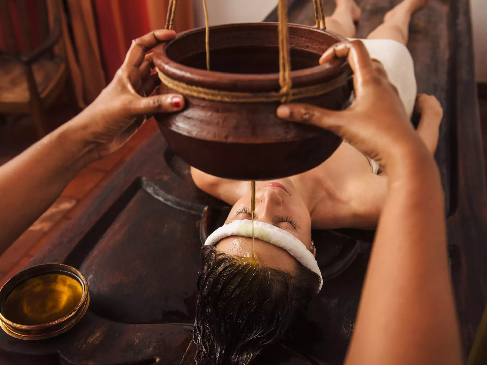 A woman lies on a treatment bed as a therapist pours a gentle stream of oil on her forehead.