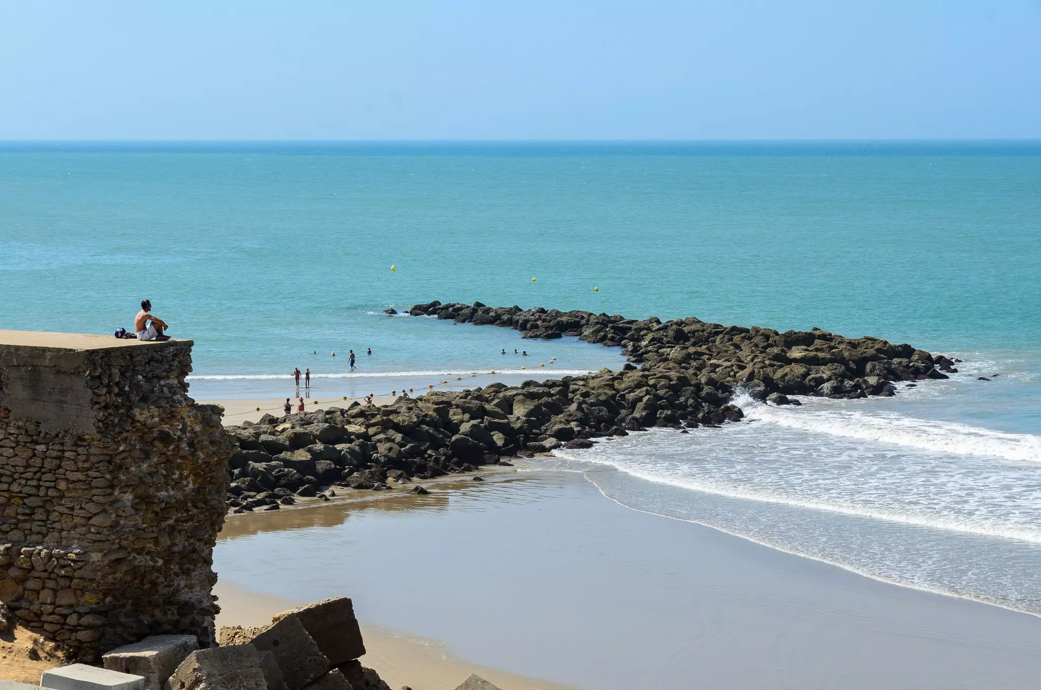 A man sits on a rocky perch overlooking a beach. People play in the water down below.