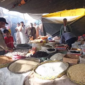 Woven baskets of grains, beans and spices at a tented market stall with shoppers behind, Had Draa, Morocco