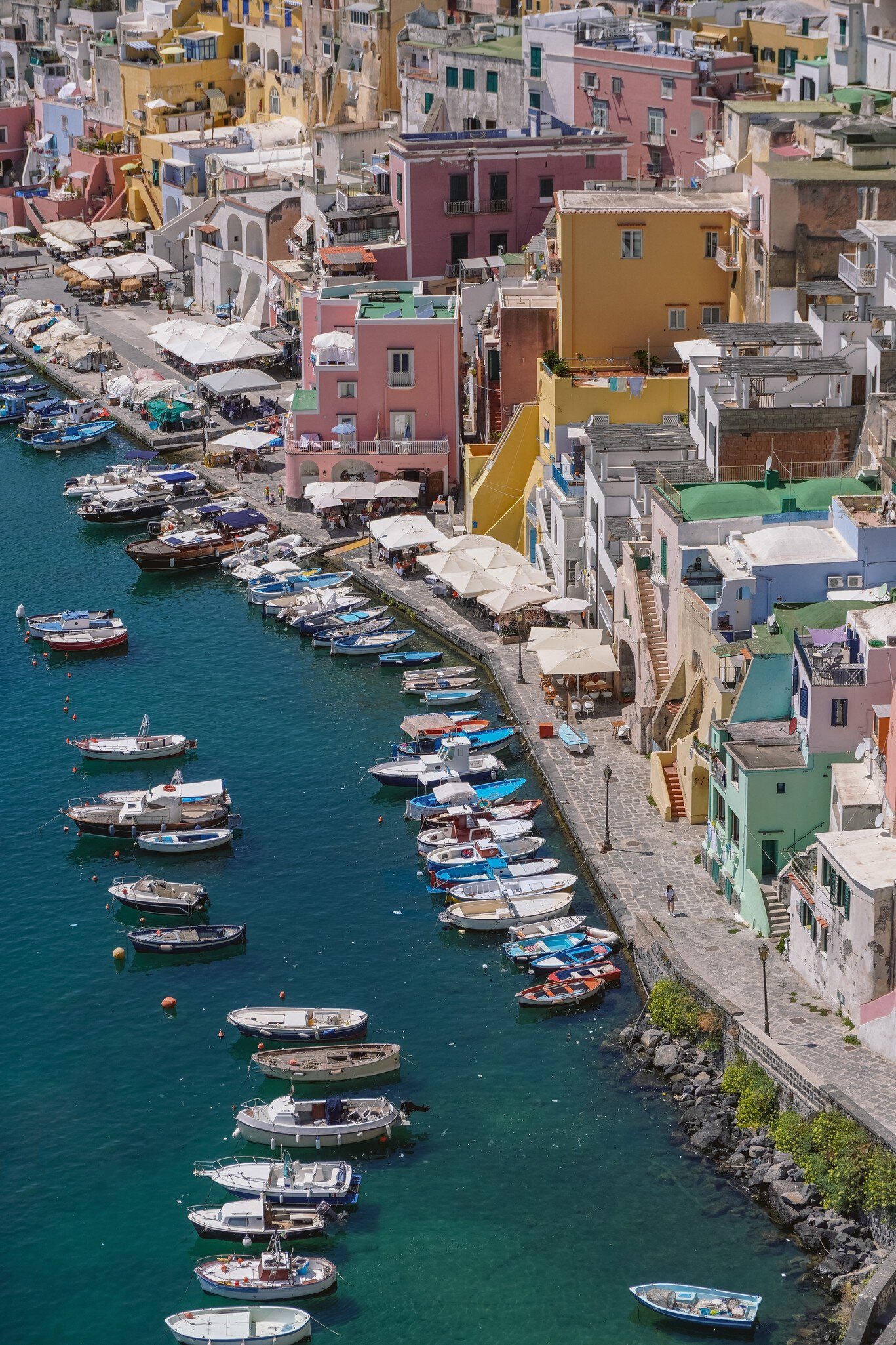 Marina di Corricella on Procida from above, with small boats moored in green water below pink, yellow and white houses.