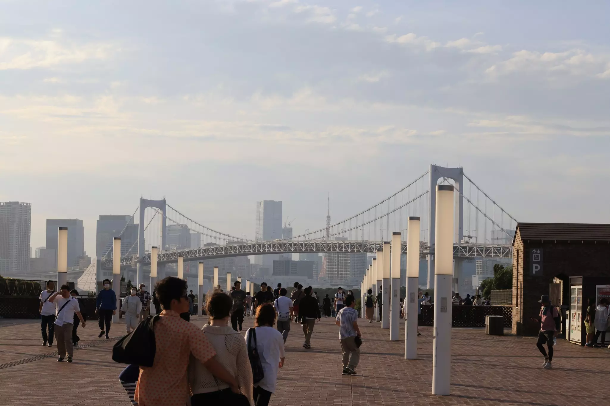 People walk on a promenade in a waterfront city park. A suspension bridge and office towers can be seen in the distance.