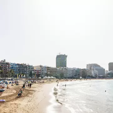 A curving city beach with pale sand and shallow turquoise water, apartment blocks and a green glass tower along the promenade.