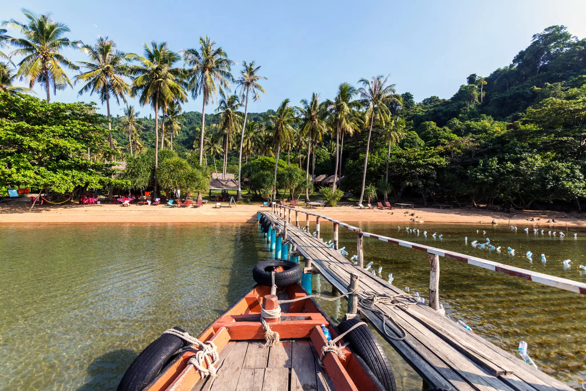 From the perspective of a boat arriving at a narrow wooden pier, leading to a sandy beach with palm trees.