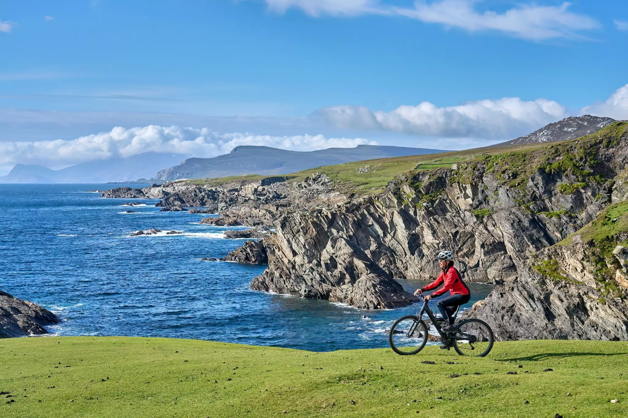 A woman cycling by sea cliffs in Ireland under a blue sky.