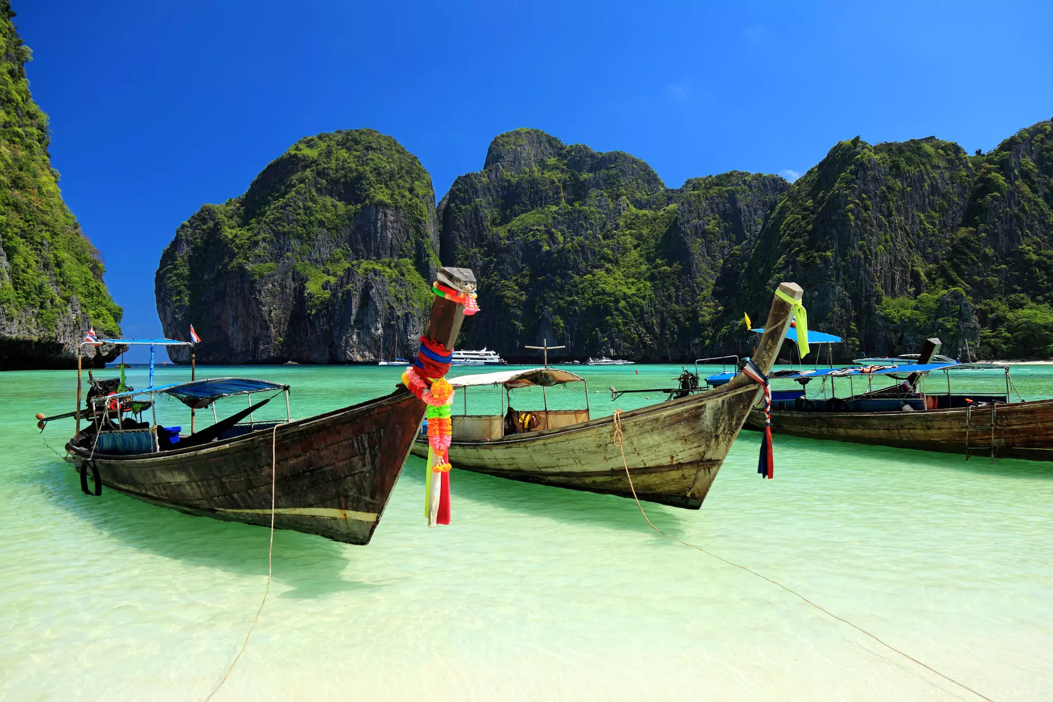 Three long-tail wooden boats with colorful ties on their bows moored in clear water on a sunny day with large rocky mountains in the distance.
