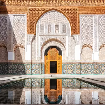 Carved stucco walls and zellige tiles surround a golden wooden door mirrored in a rectangular pool of still water