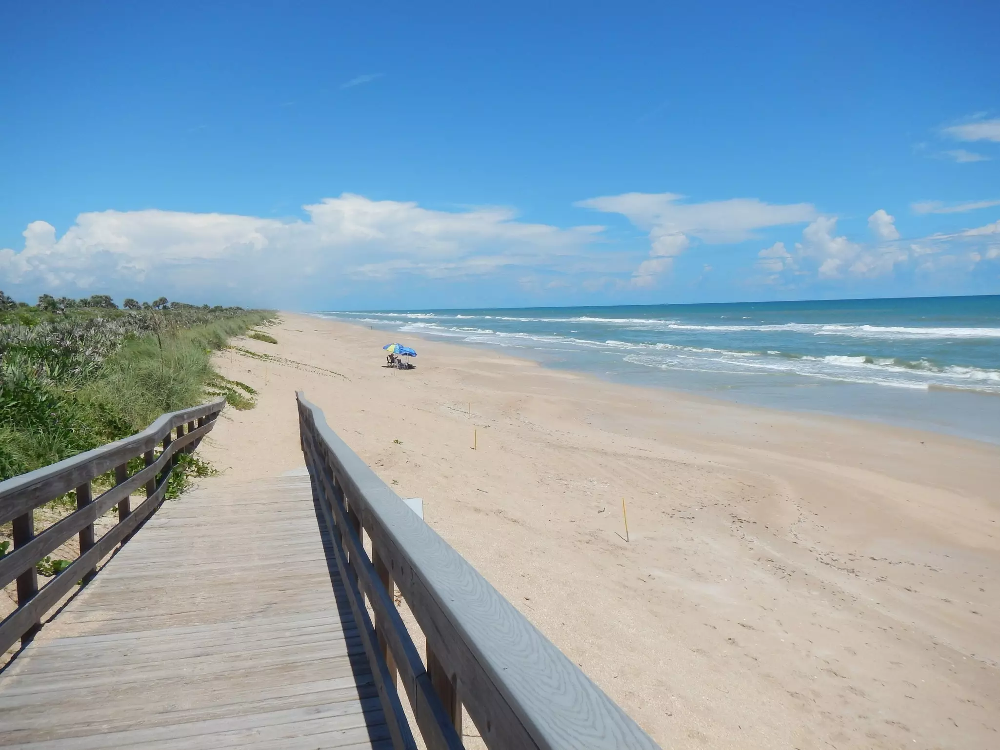 A boardwalk leads down to a long sandy white-sand beach with just two people who have set up camp under blue and yellow sunshades.