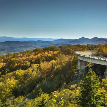 An RV drives toward a curve on a raised roadway above trees in warm light; the silhouette of a mountain range is in the background.