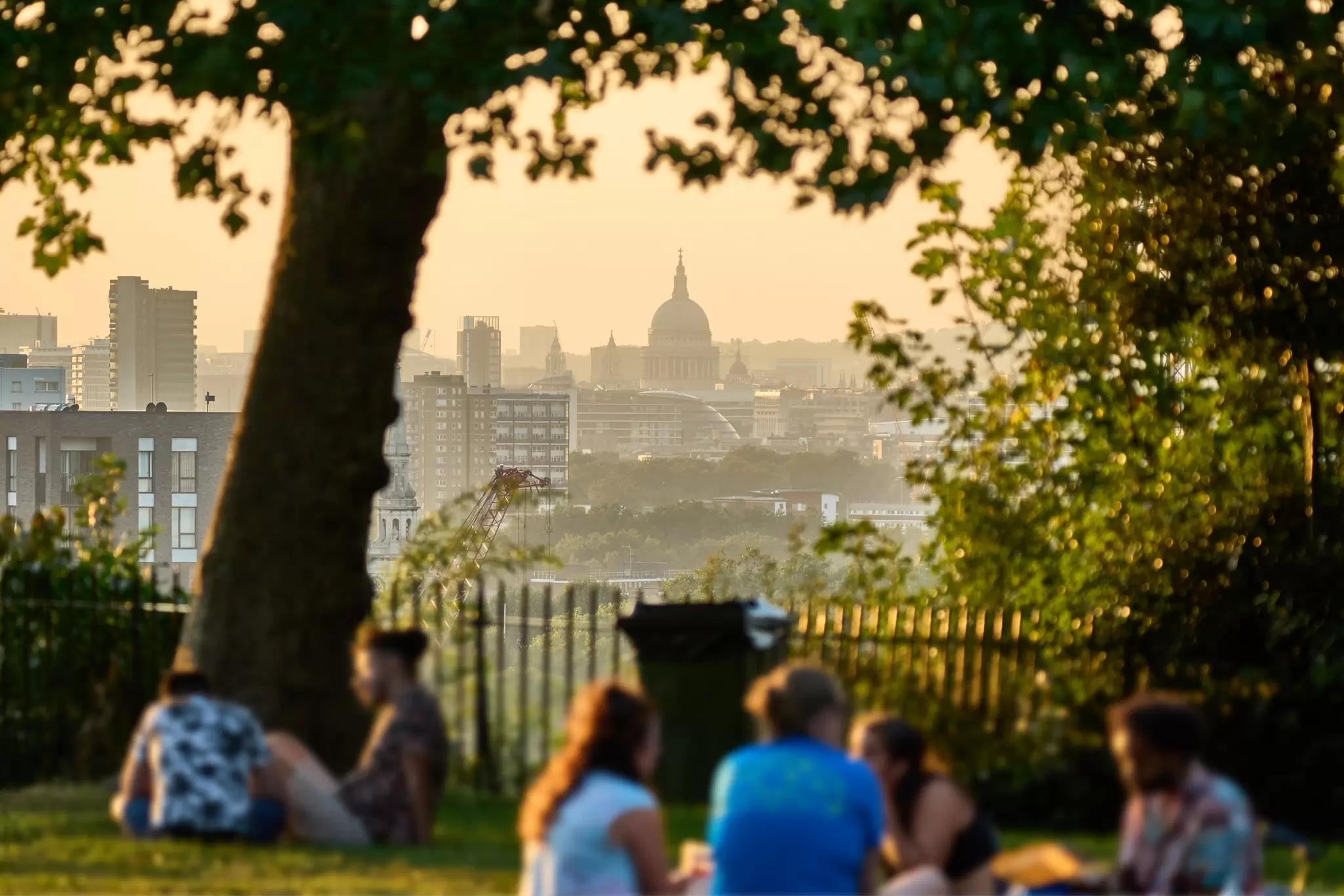 People gather at a small garden viewpoint in a city, with views of a domed church in the distance: St Paul's Cathedral viewed from The Point in Greenwich. 