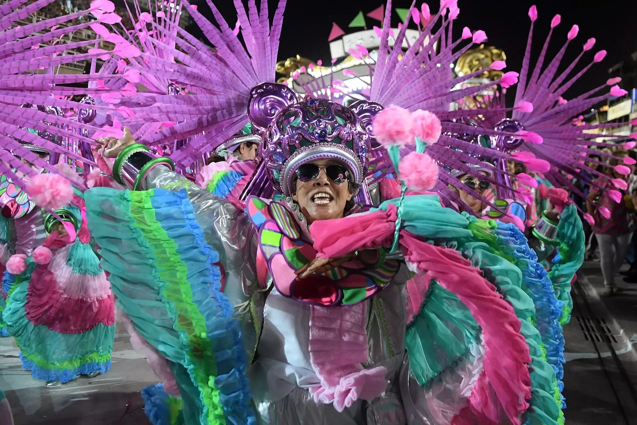 A Carnaval participant wearing a pink, green and blue costume while smiling and standing in front of pink and purple feather accessories