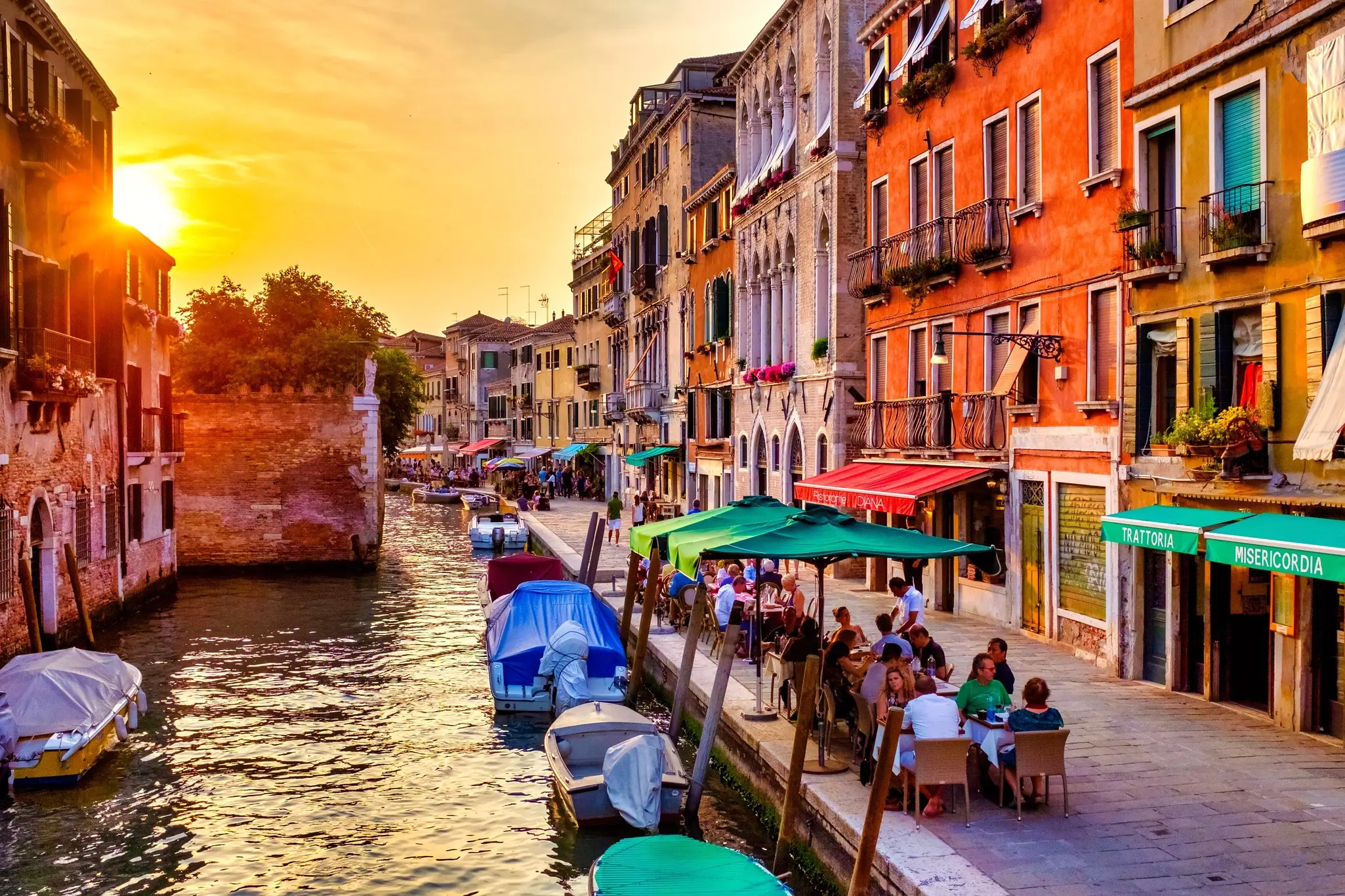 Diners sit at restaurant tables beside a canal as the sun sets casting an orange glow over the buildings.