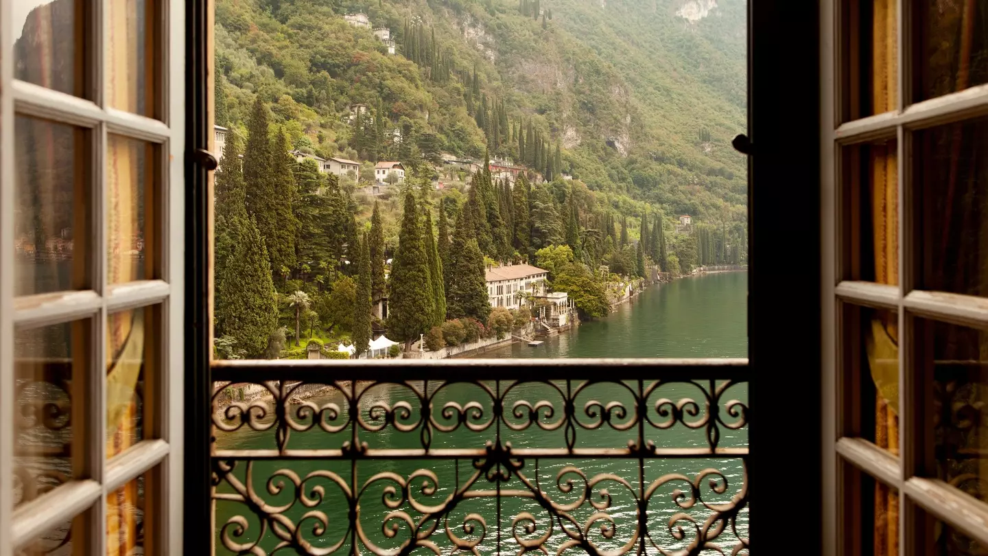 A lake and houses beyond iare seen through an open window, which has an ornate metal railing.