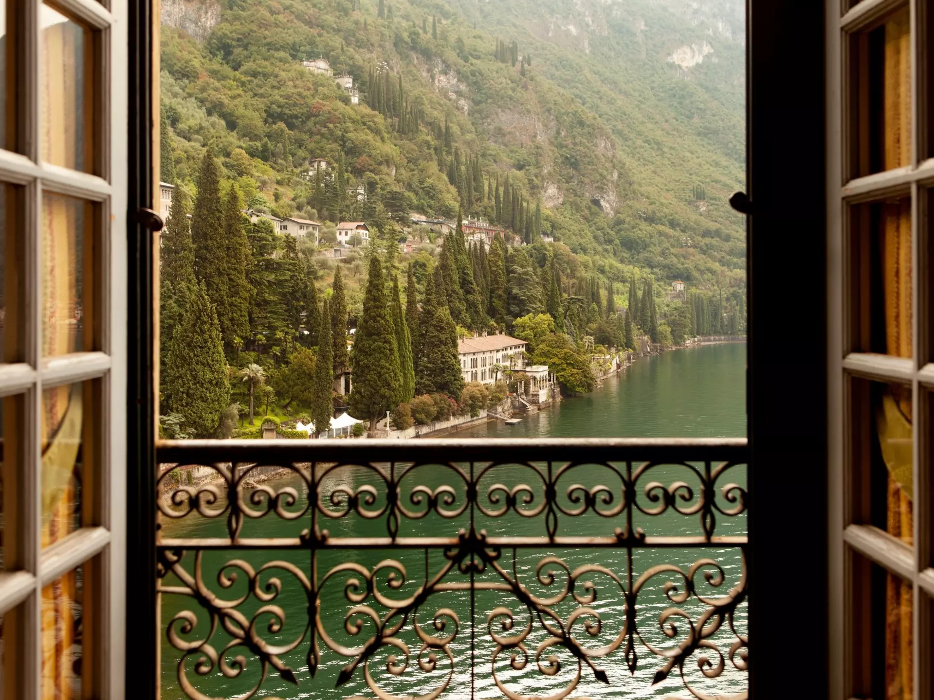 A lake and houses beyond iare seen through an open window, which has an ornate metal railing.
