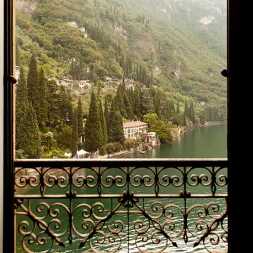 A lake and houses beyond iare seen through an open window, which has an ornate metal railing.