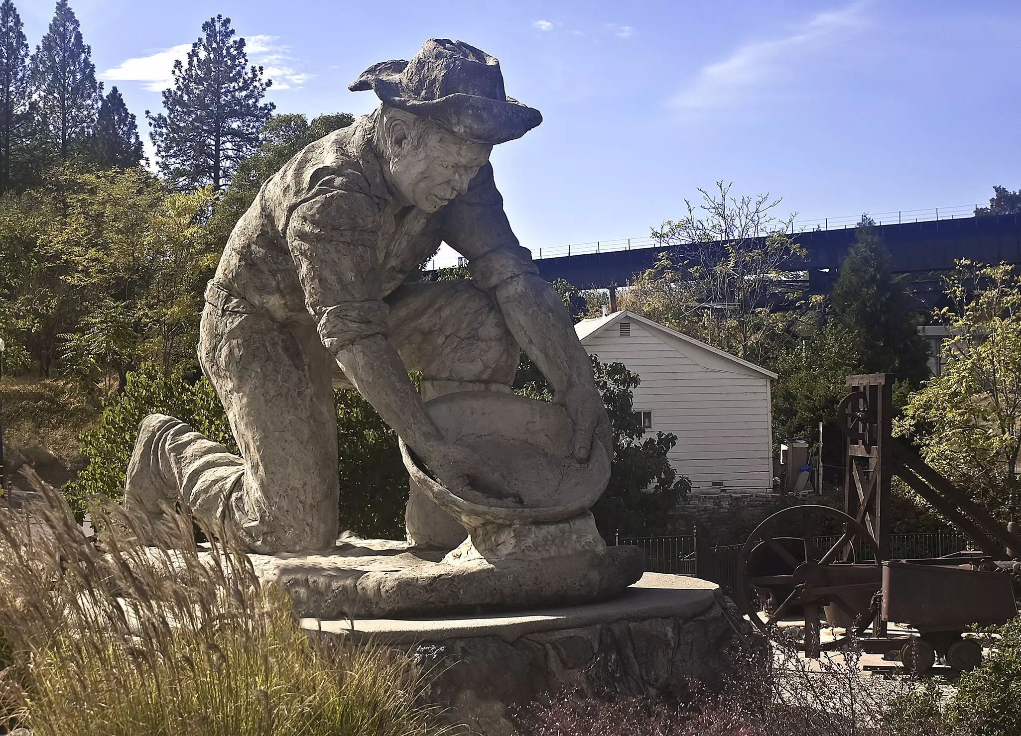 Large concrete statue of gold miner leaning over with gold pan. Old mining equipment and a raised bridge are in the background.