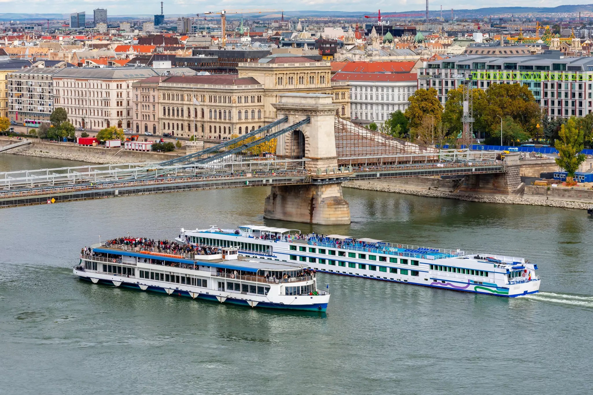 Two passenger ships pass one another in a river, under the span of a historic bridge. Buildings of the city are seen on the shore.