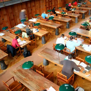 Overhead view of a wood-panelled library reading room with green desk lamps and people studying.
