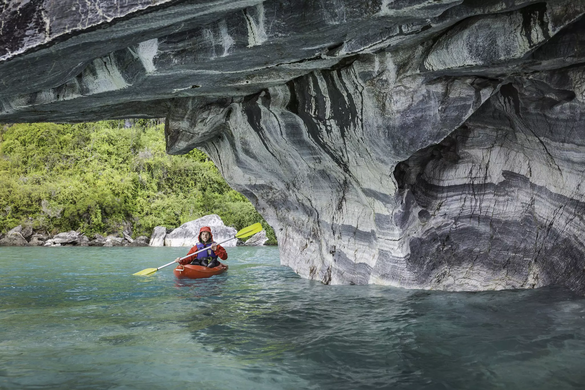 A kayaker leaves turquoise waters and enters a cave with grey and white streaked rocks.