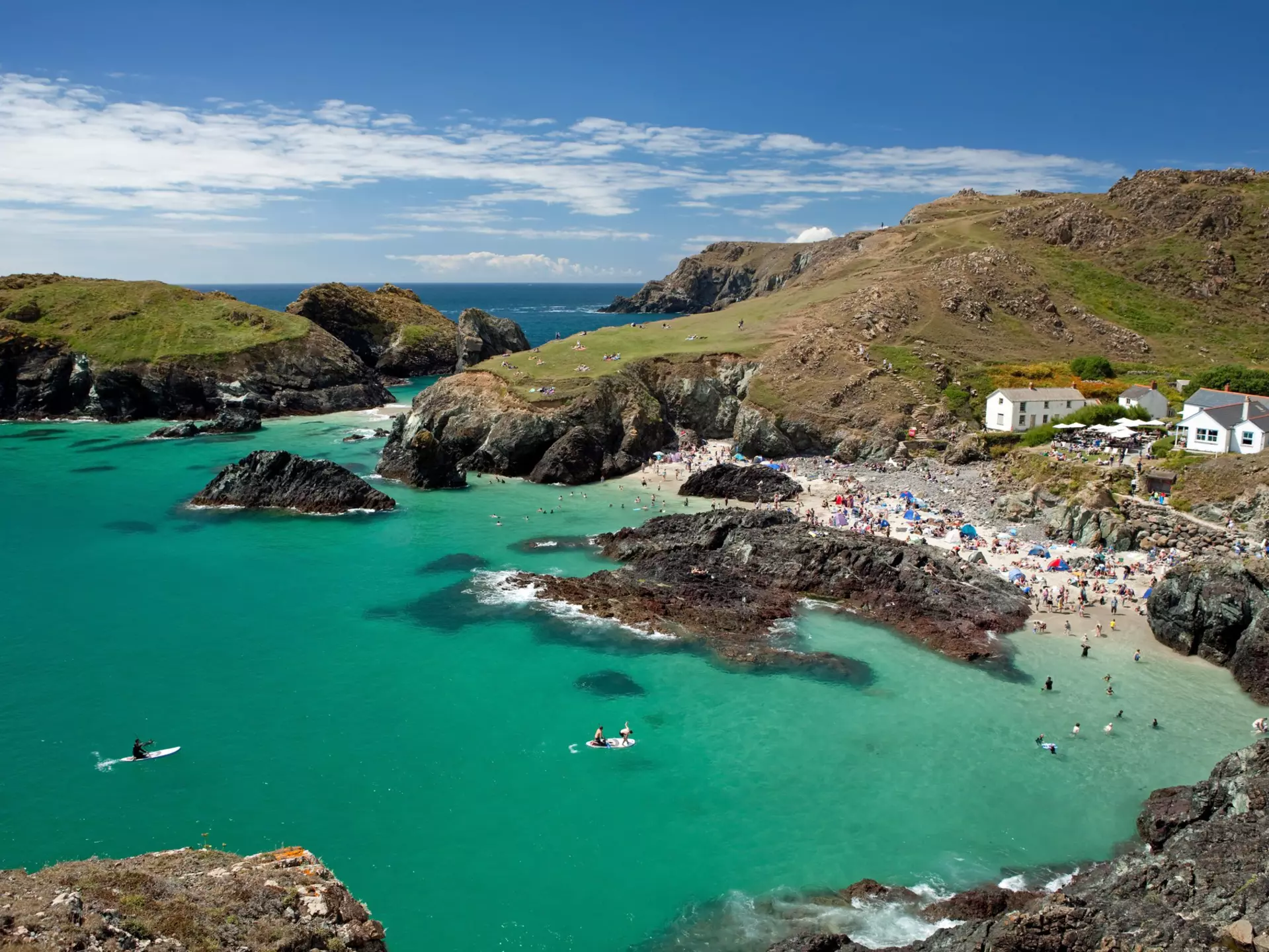 People fill a tiny sandy cove sheltered between cliffs. A white cottage cafe stands on the cliff.