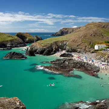 People fill a tiny sandy cove sheltered between cliffs. A white cottage cafe stands on the cliff.