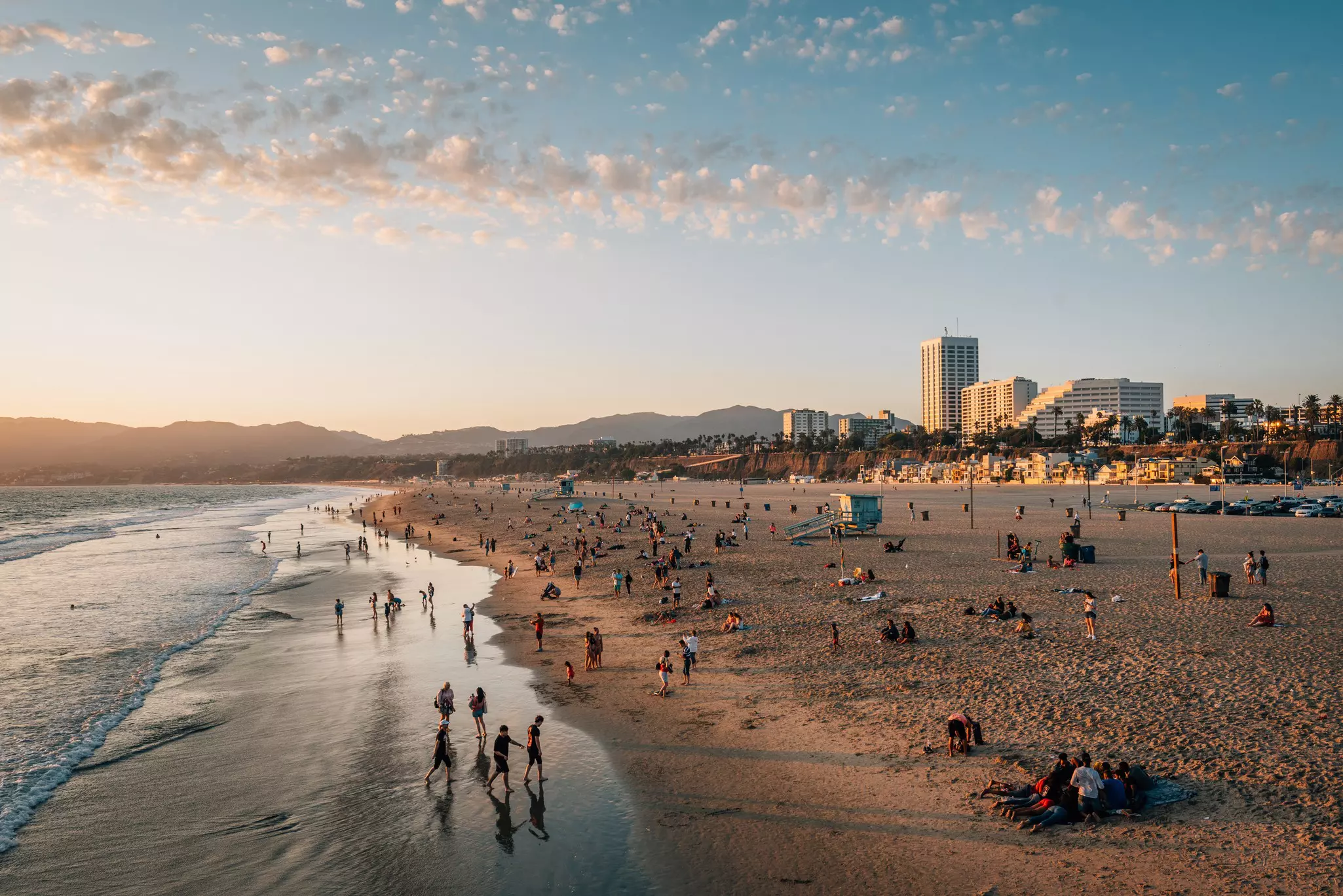 Beach at sunset looking towards the mountains. Waves lapping on a shore covered in humans with tall white buildings on the right side.