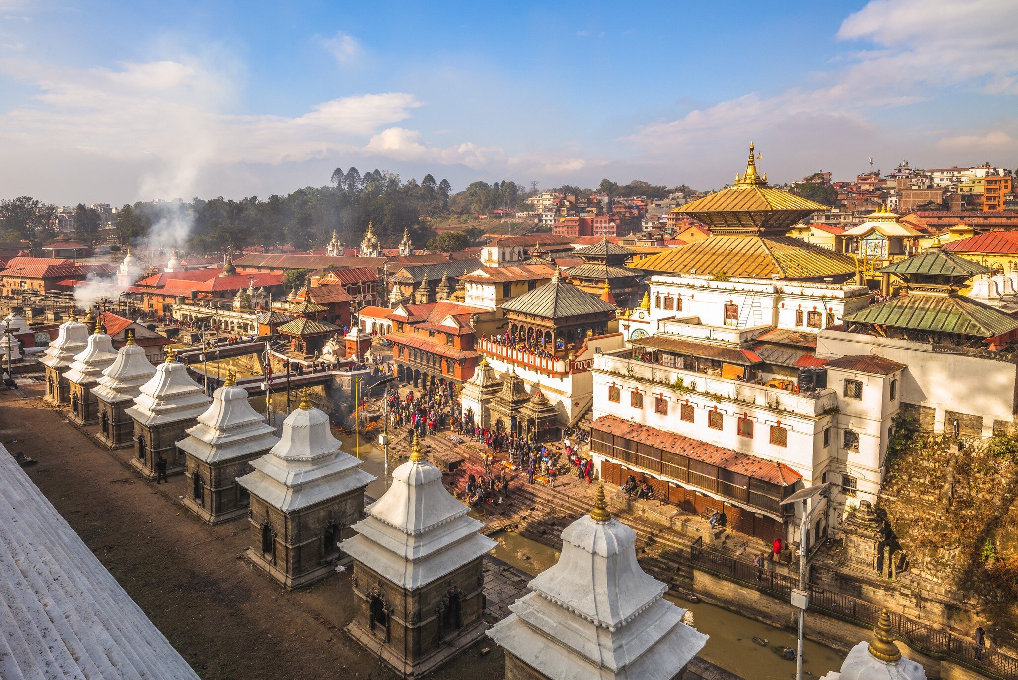 Row of white stone shrines facing a gilded two-tiered roof, with crowds on riverside steps and drifting smoke