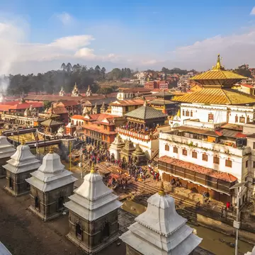 Row of white stone shrines facing a gilded two-tiered roof, with crowds on riverside steps and drifting smoke