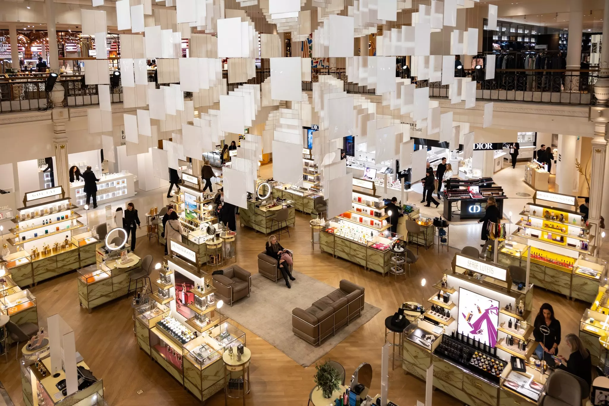 An overhead view of kiosks selling cosmetics at a department store, under suspended panels in an atrium.