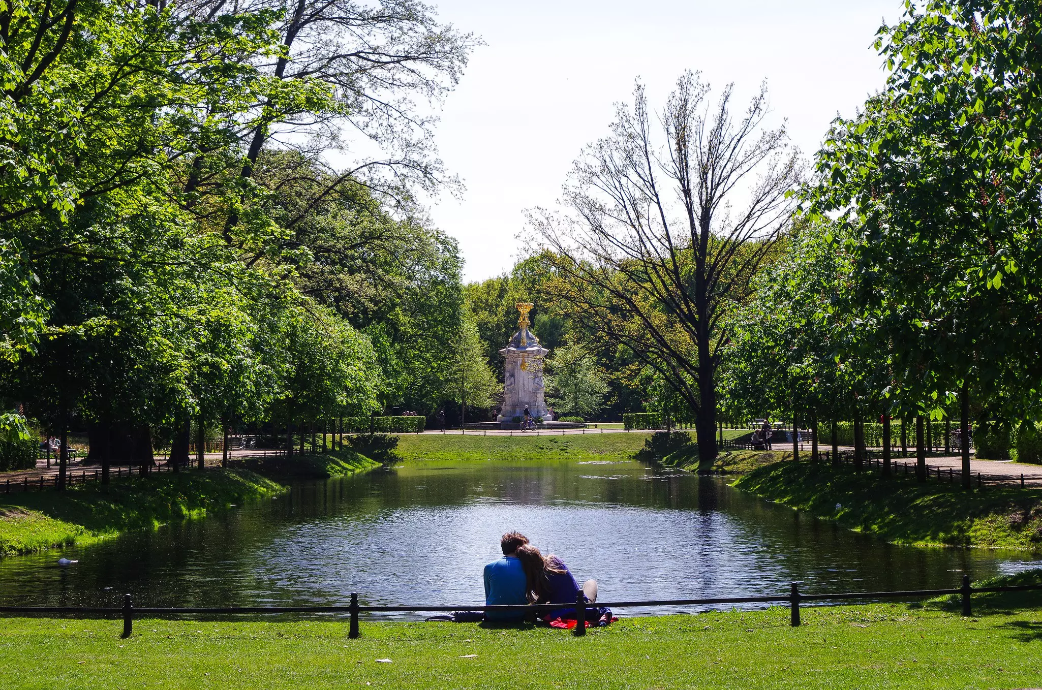 A couple sat together by a pond in green parkland.