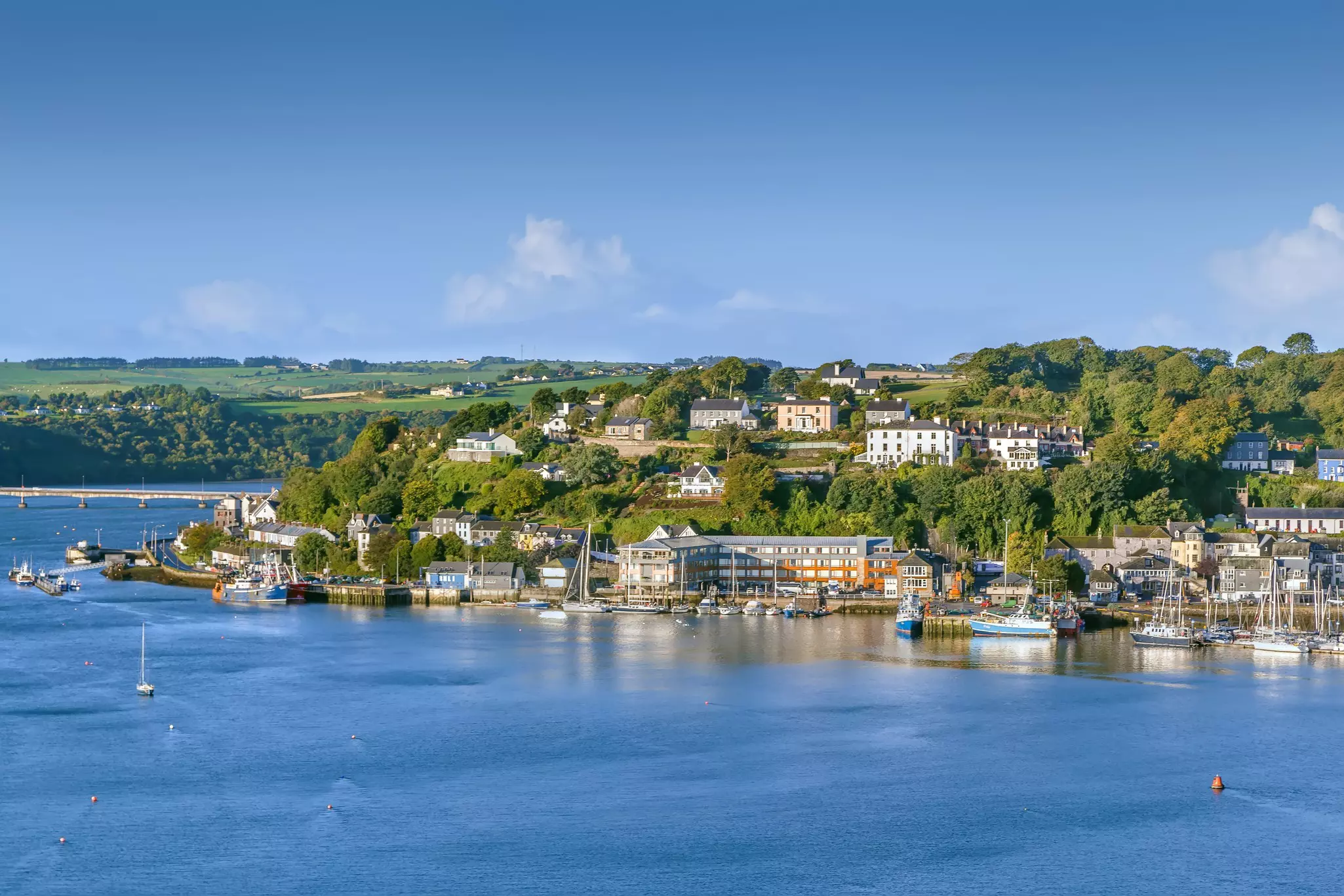 The low buildings of a small coastal town in Ireland face blue water; there are green fields in the background.