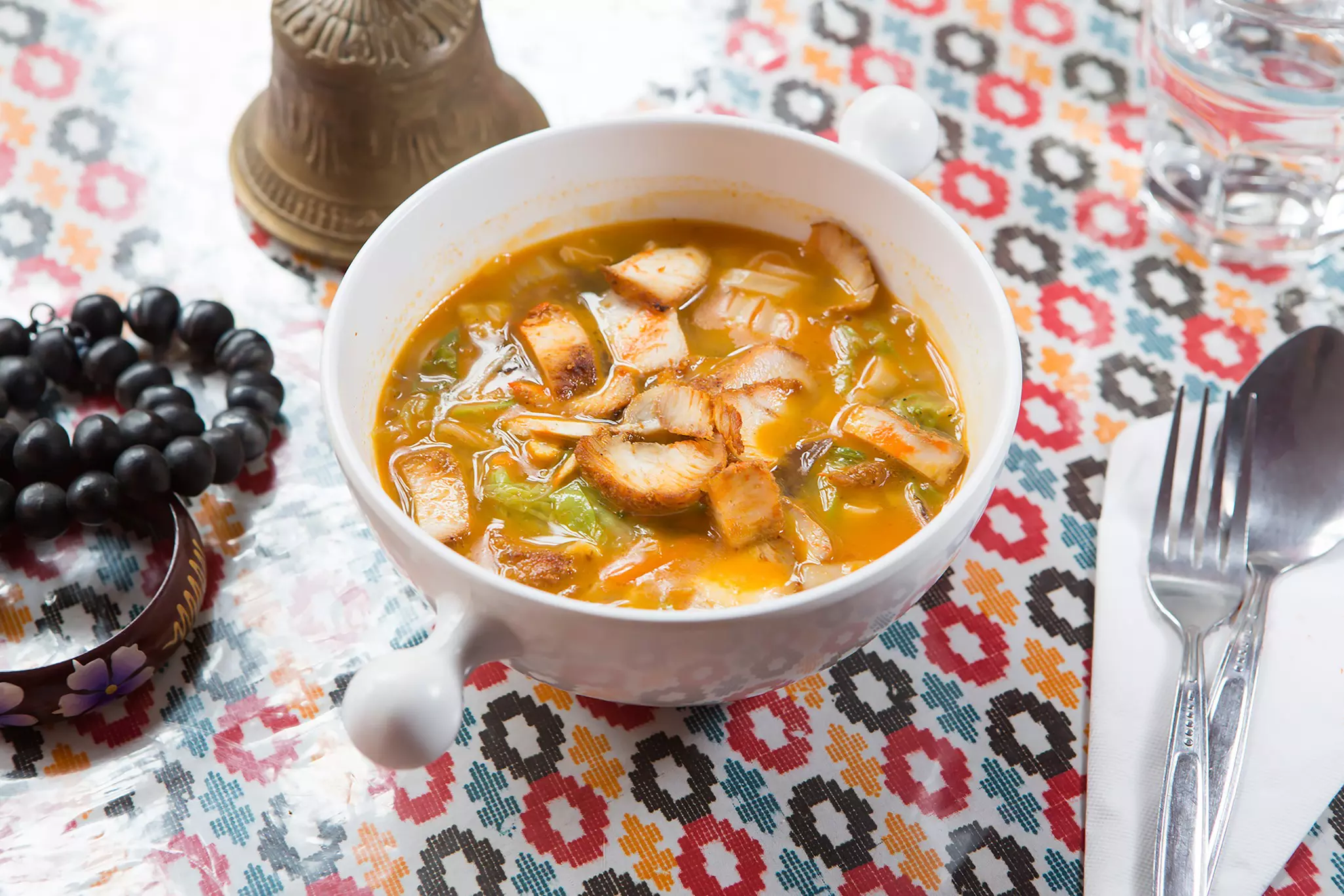 Thukpa, a bowl of noodles with meat, vegetables and broth on a table with cutlery, prayer beads and a bell.