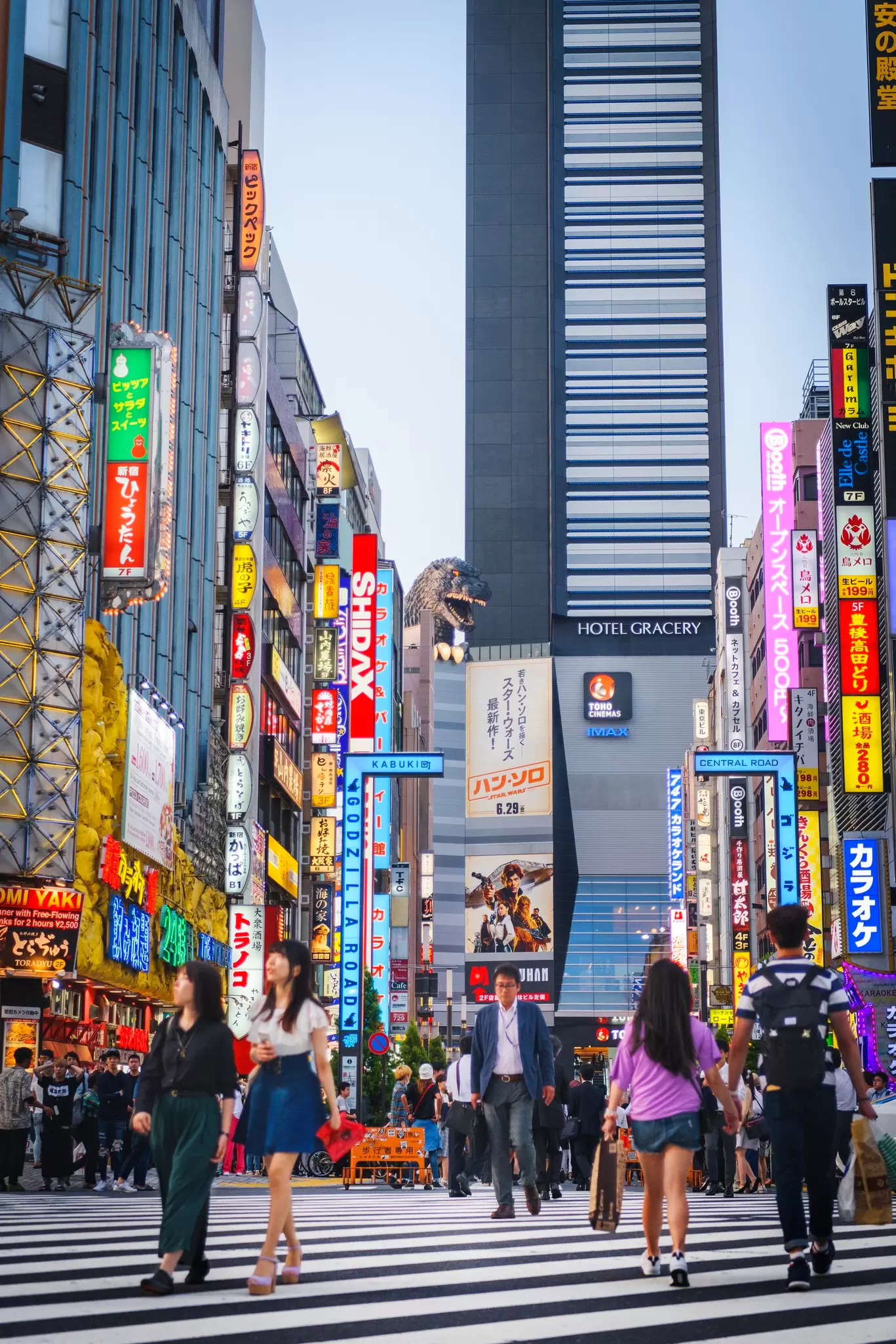 A busy intersection with neon signs; at the far end is a hotel with a large Godzilla statue.