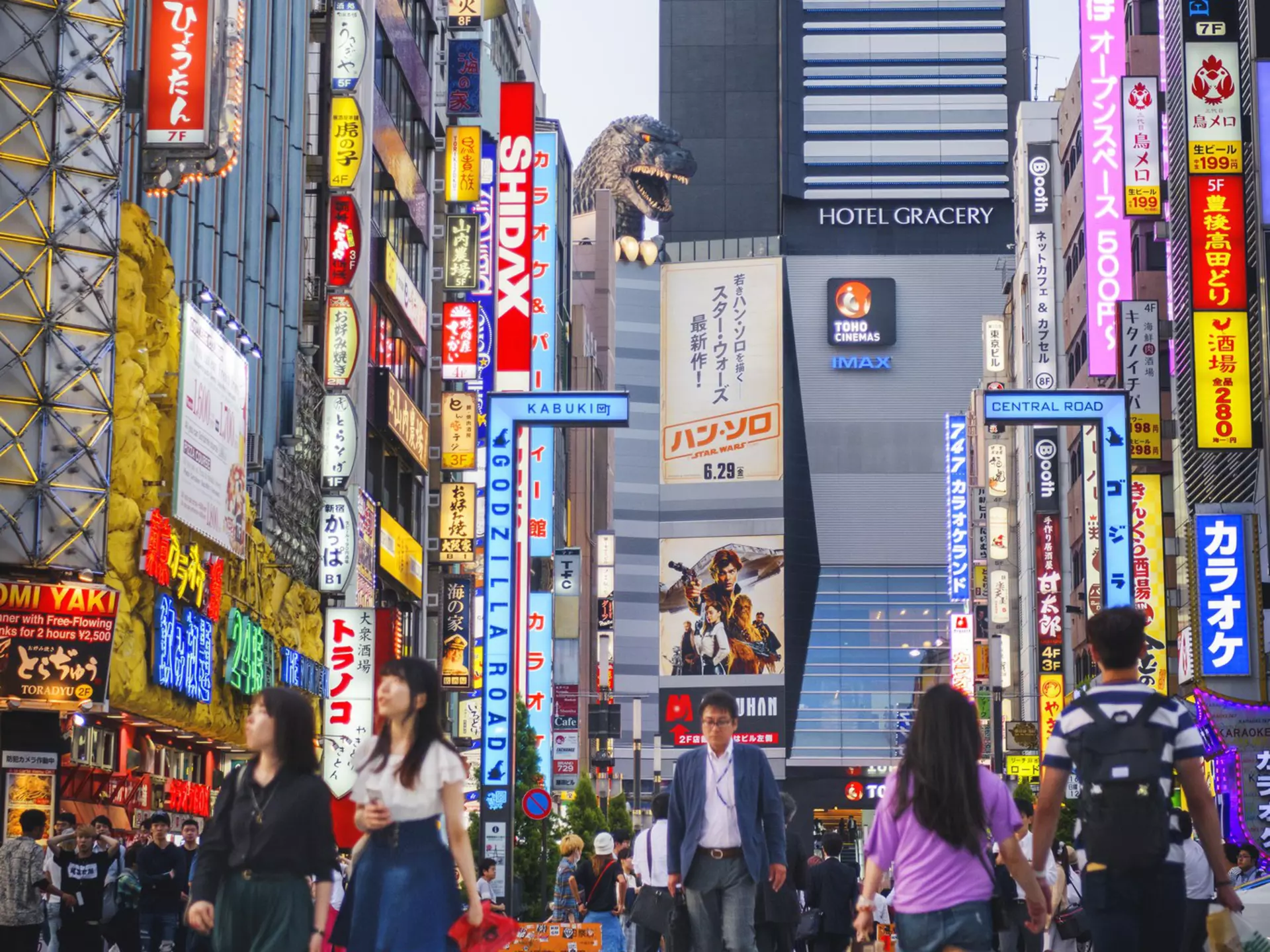A busy intersection with neon signs; at the far end is a hotel with a large Godzilla statue.