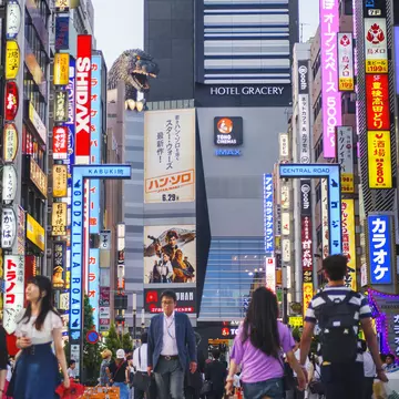 A busy intersection with neon signs; at the far end is a hotel with a large Godzilla statue.