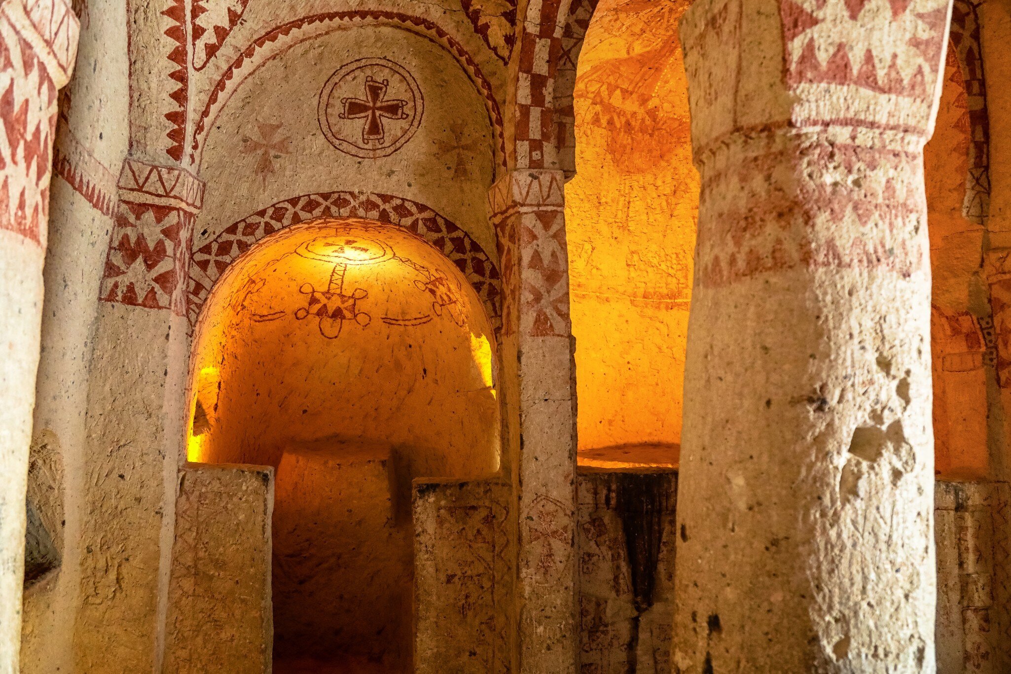 Rock-cut chapel interior painted with red geometric patterns and crosses, its apse lit against pale stone.
