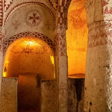 The interior of a rock-cut chapel painted with red geometric patterns and crosses, its apse lit warm against pale carved stone.