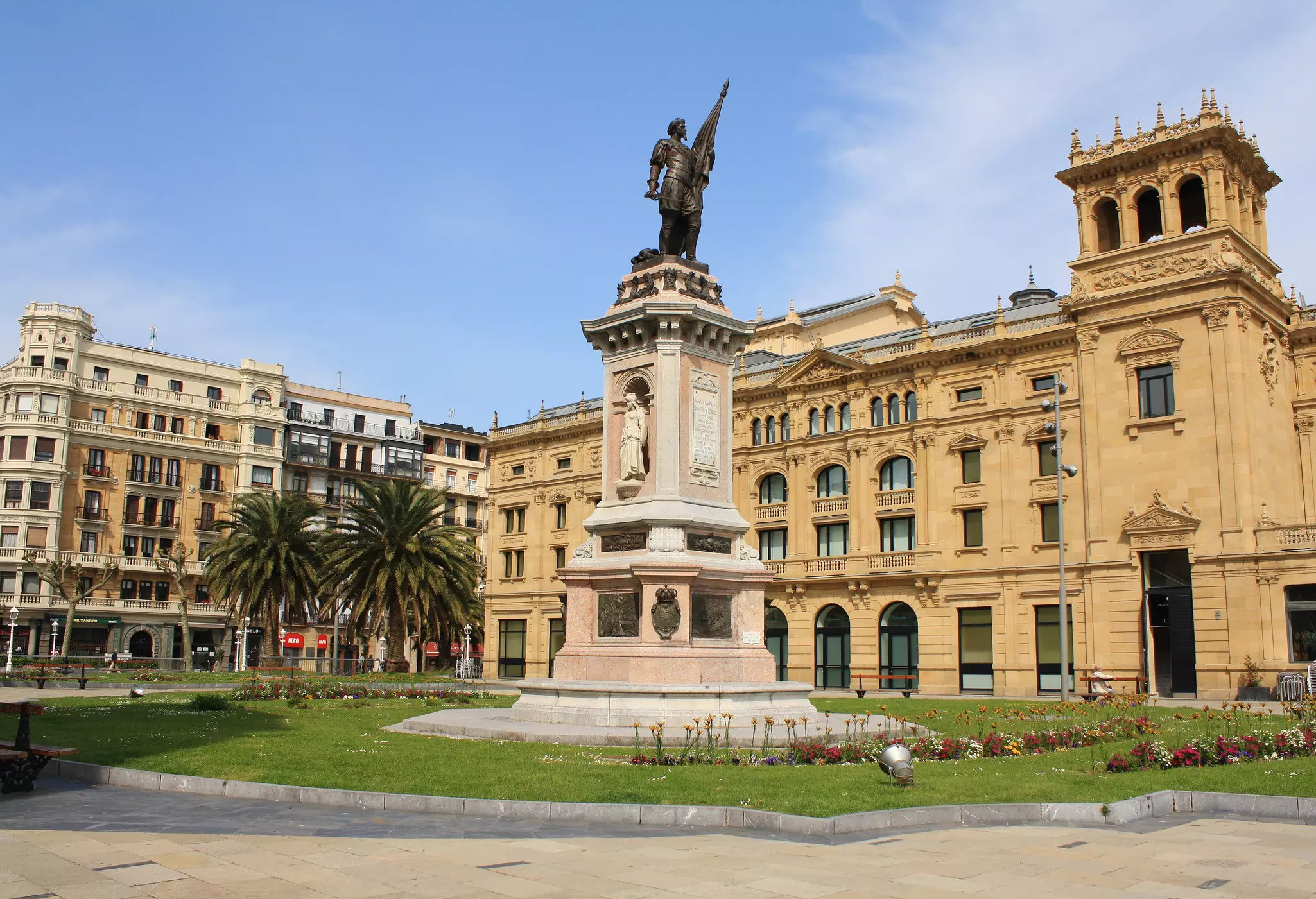 The exterior of a grand theater building with ornate stonework. A statue on a tall plinth stands in gardens at the front.