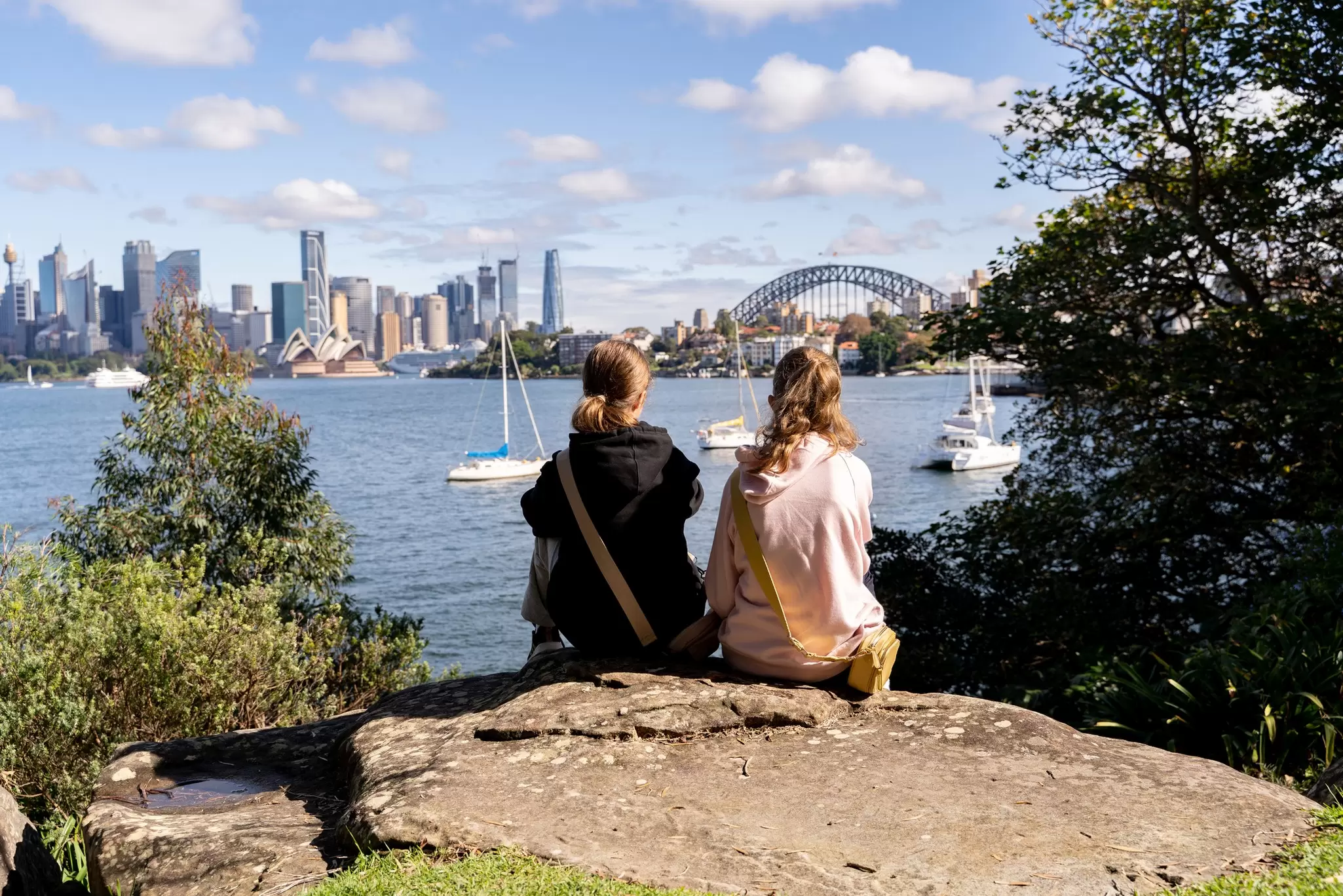Two girls sit on a rock and look out over an iconic harbor, with a white opera house and a large bridge