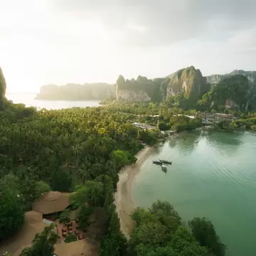 Palm-covered peninsula with a curved sandy beach, moored longtail boats and tall limestone cliffs in hazy light.