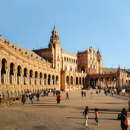 Tourists stroll a semi-circular plaza lined with a mix of Renaissance and Moorish architectural styles, adorned with colorful ceramic tiles.