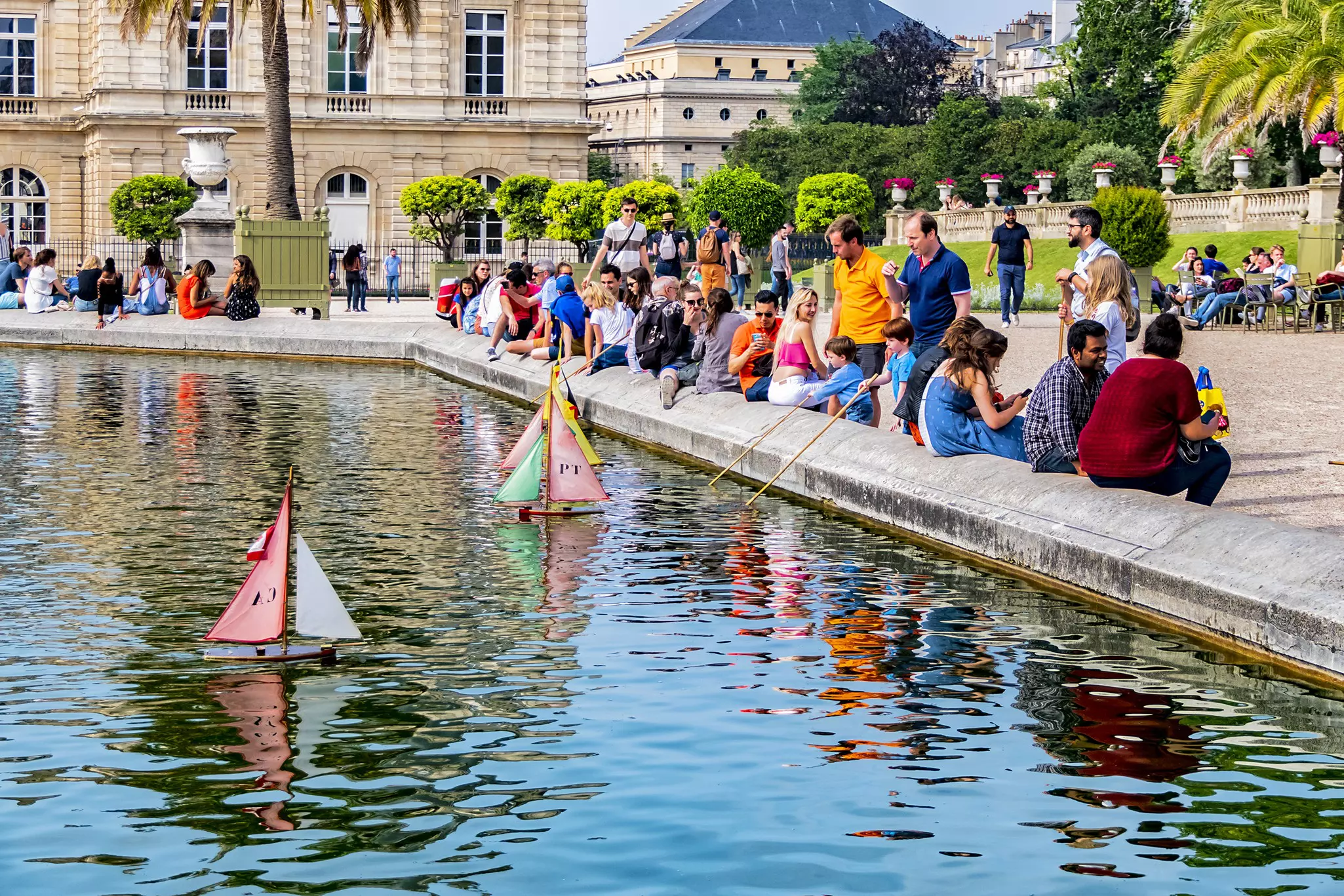 People stand at the ledge of a pond in a park as they operate toy boats on the water. An ornate palace is visible in the background.