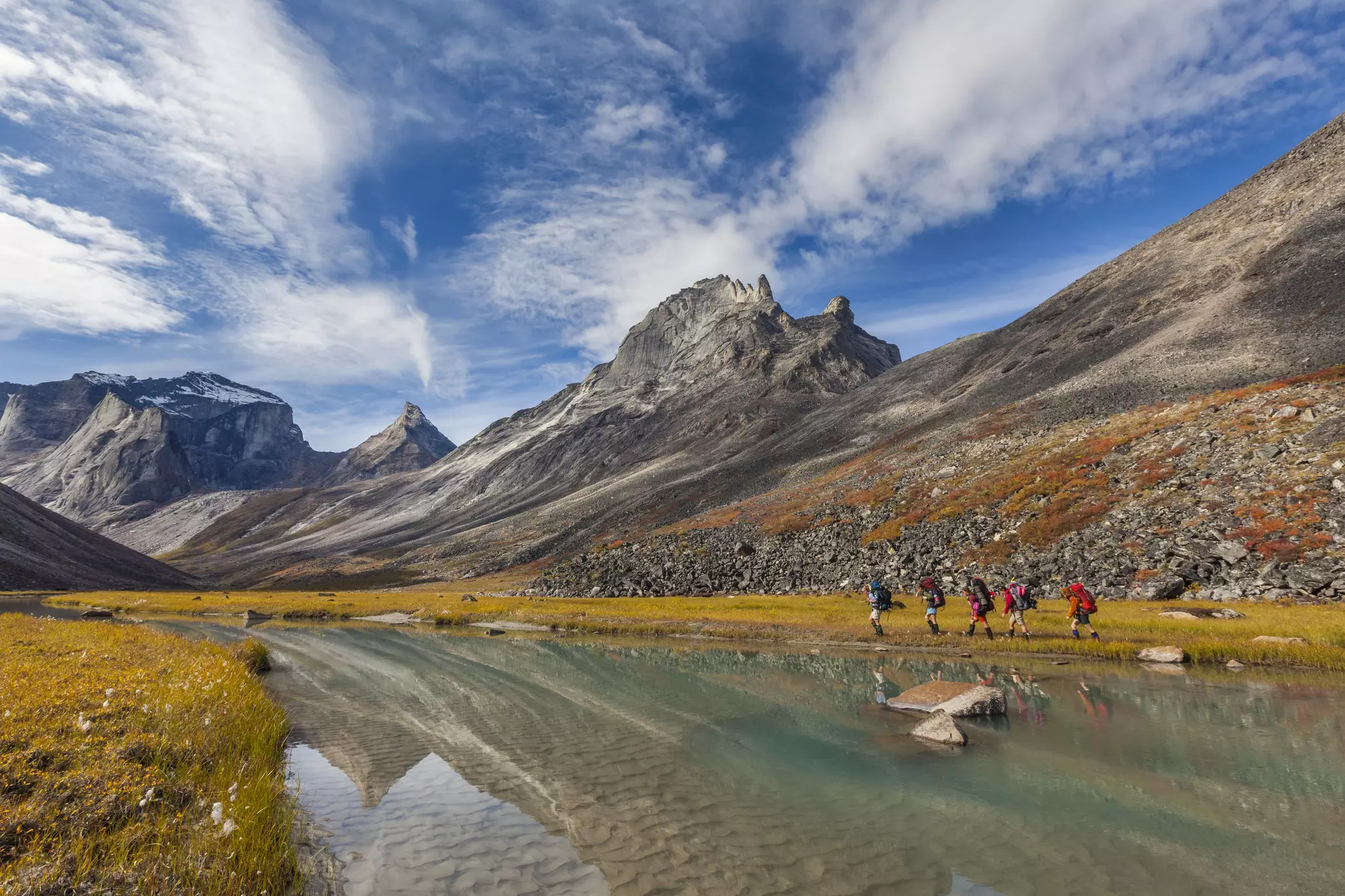 Backpackers walk along the shore of a creek, with rocky peaks in the background.