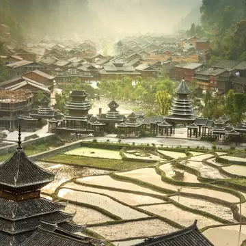 Dark-tiled wooden houses and tiered drum towers beside water-filled rice terraces in Zhaoxing, China, under morning mist.